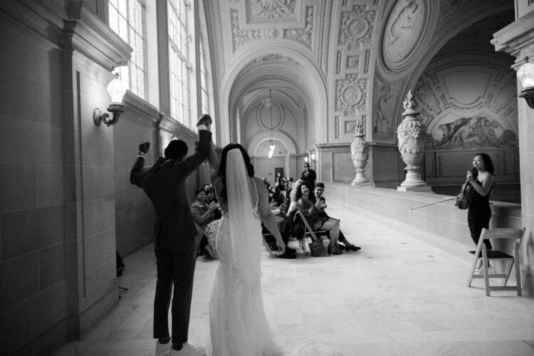 A shot of the couple raising their hands in joy after the ceremony captured by a San Francisco City Hall wedding photographer on the 4th floor balcony