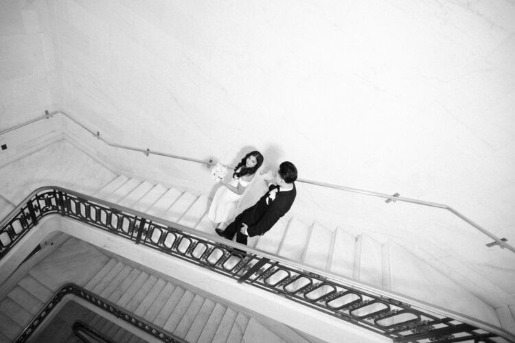 A couple going down the spiral staircase at city hall. The photo is black and white and looks like a classic film picture. captured by a San Francisco City Hall wedding photographer